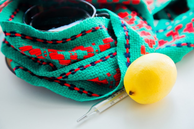 A cup of tea with lemon stands on a knitted green and red scarf there is a thermometer and a lemon n...