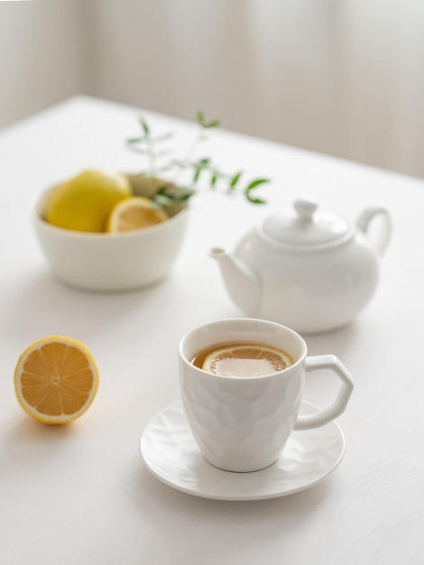 A cup of tea with lemon and a teapot on a white table against the background of a kitchen window