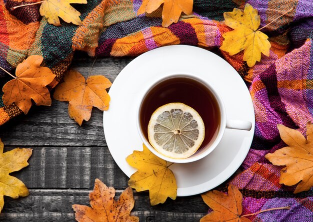 Cup of tea with lemon with scarf and autumn leaves on wooden surface