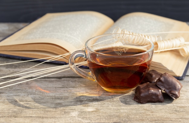 Cup of tea with open book and chocolate sweets on wooden table.