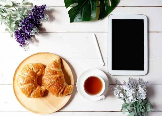 Cup of tea with tablet, green leaf, flower and wooden dish with croissants on white wooden background.