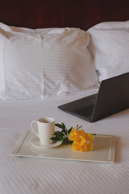 Cup of tea with a yellow flower on a white bed next to laptop.