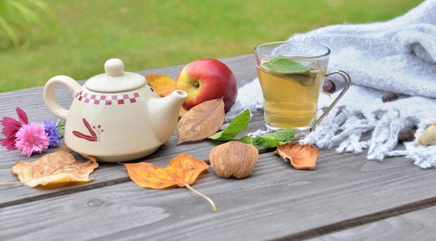 Чашка чая на деревянном столе в саду с чайником среди осенних листьев и красного яблока на шерстяном шарфе