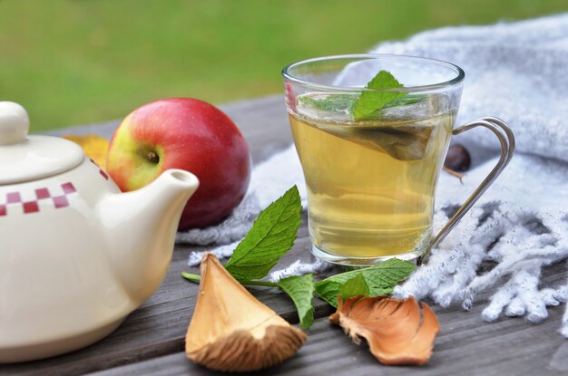 Cup of tea  on a wooden table in garden with teapot  and red apple on wool scarf