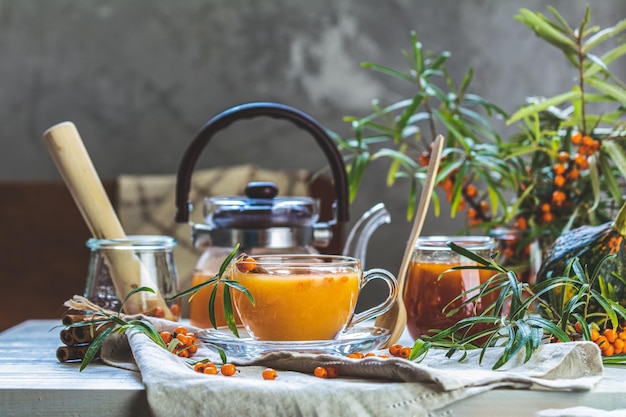 Cup and teapot of hot spicy tea with sea buckthorn jam in the glass jar branches of fresh berries on light woden table surface in the rustic room
