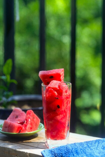 A cup of watermelon juice and sliced watermelon are put on the\
wooden board of the balcony