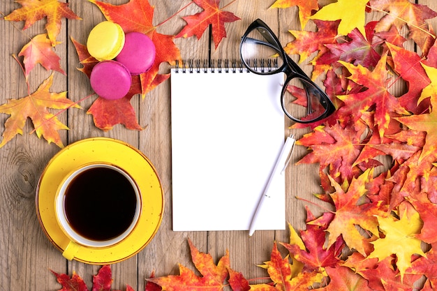 Photo cup with black coffee, glasses, yellow lollipops, macaroons, notepad, wooden table with autumn fallen orange leaves flat lay