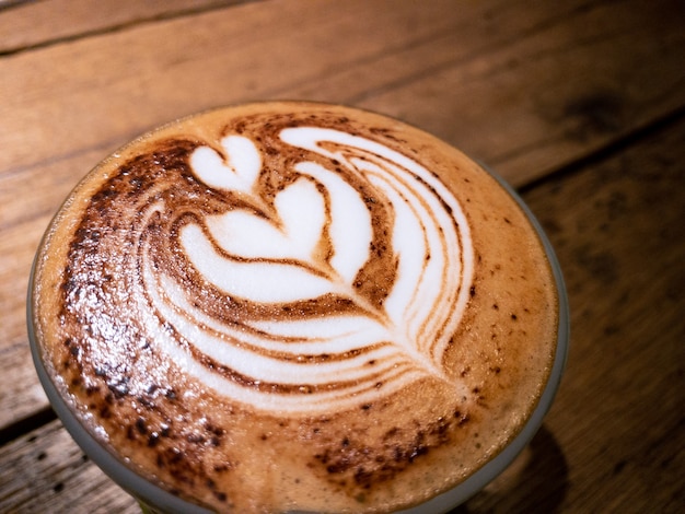 Чашка с капучино с пеной искусства латте в форме сердца на черном деревянном столе в кофейне. Дымящийся горячий кофеиновый напиток на столе в кафе.