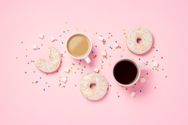 Cup with coffee or tea, Fresh tasty sweet donuts, colorful decorative candies, sugar cubes on a pink background. Bakery concept, fresh pastries, delicious breakfast, fast food. Flat lay, top view.