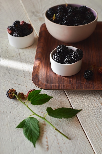 cup with fresh black and red blackberries on a brown tray, green blackberry leaves is on the table. Light wood background