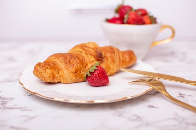 Cup with fresh strawberry and tasty croissant on white plate