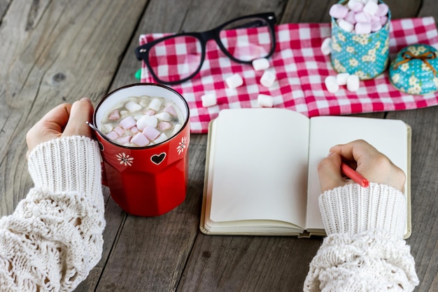 A cup with marshmallows and a pen for writing in female hands at the table