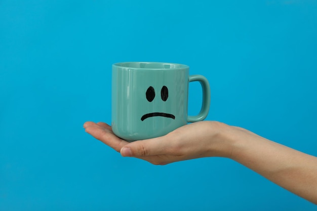 Cup with sad emoji in hand on blue background