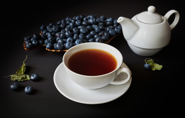 cup with tea, teapot, blueberry and dry leaves of mellisa on dark background