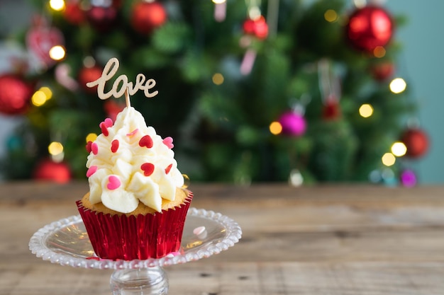 ディスプレイのカップケーキは、LOVEサインと背景の焦点が合っていないクリスマスツリーと木製の背景に立っていますコピースペース