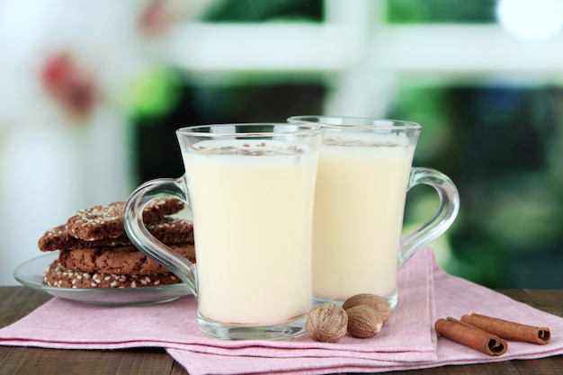 Cups of eggnog with cinnamon and cookies on table on bright background