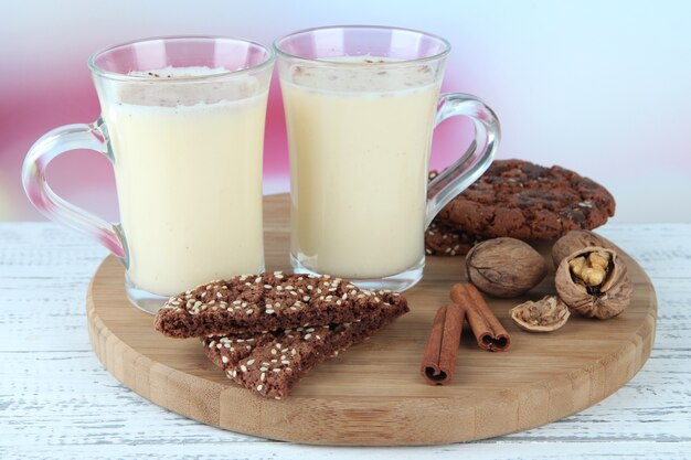 Cups of eggnog with cinnamon and cookies on table on bright background