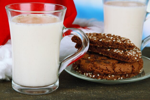 Cups of eggnog with cookies and santa hat on table on bright background