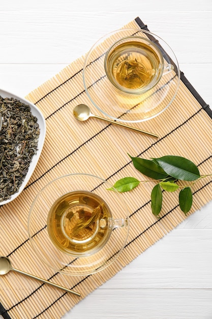 Cups of green tea and dry leaves on bamboo mat