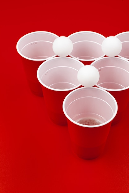 Cups and plastic ball on red background. Beer pong game