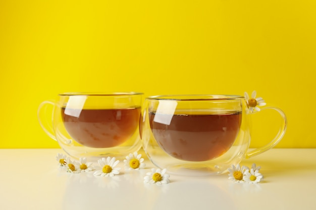 Cups of tea and chamomile on white table against yellow background