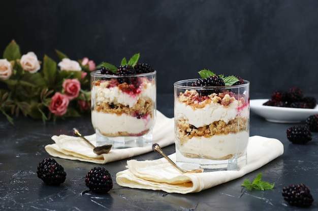 Curd dessert with blackberries and cookies in glass on a dark table