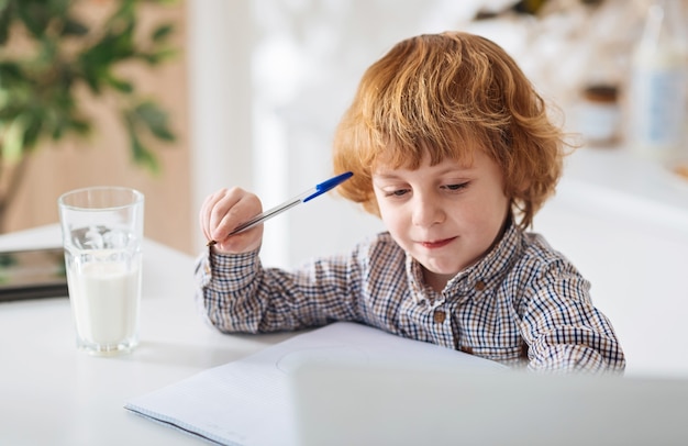 Curiosity is the key. Lovely admirable smart child sitting at the table in the kitchen and writing down notes while drinking a glass of milk