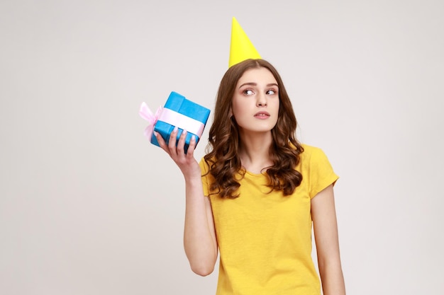 Curious adorable woman of young age in yellow casual Tshirt shaking blue giftbox near ear trying to guess her present expectating expression Indoor studio shot isolated on gray background