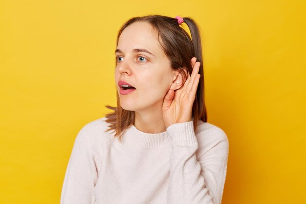 Любопытная внимательная девочка-подросток в свитере с хвостиками пытается подслушать, как кто-то держит руку возле уха, пытается понять изолированное на желтом фоне