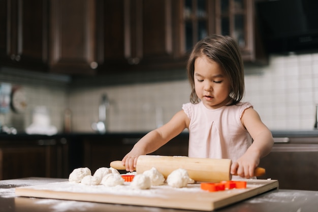 Curious beautiful little girl is rolling the dough out with wooden rolling pin at the table