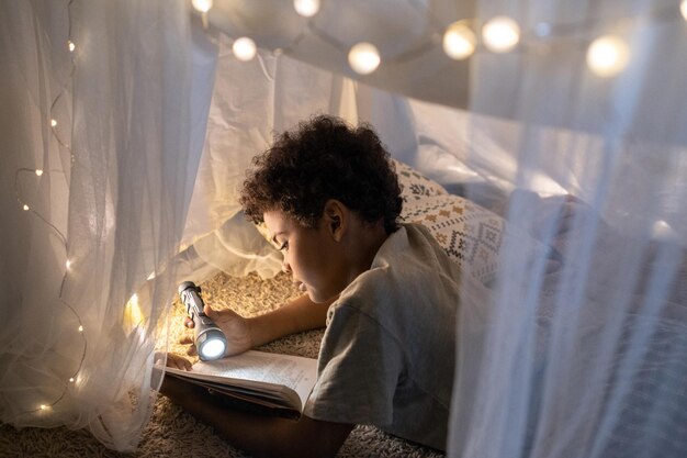 Curious black boy with curly hair lying inside of play tent decorated with christmas lights