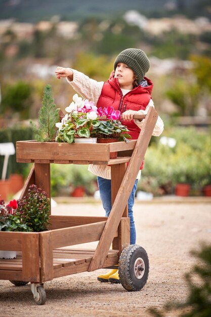 Curious boy near wooden cart with potted flowers pointing\
away