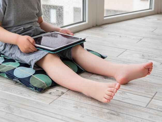 Foto ragazzo curioso guarda cartoni animati su tablet digitali ragazzino si siede sul pavimento e usa dispositivi elettronici all'interno