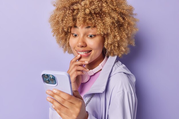 写真 スマートフォンの画面に焦点を当てた好奇心旺盛な陽気な巻き毛の女性は、エキサイティングなニュースを読みます唇は紫色の背景に隔離されたジャケットに身を包んだ驚きの表情をしています人々の反応技術