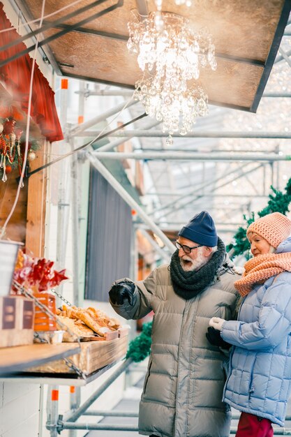 Curious couple of pensioners smiling while standing in front of the counter at the fair in winter