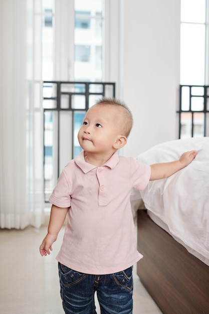 Фото Любопытный милый маленький мальчик, стоящий на кровати, трогая одеяло и глядя вверх