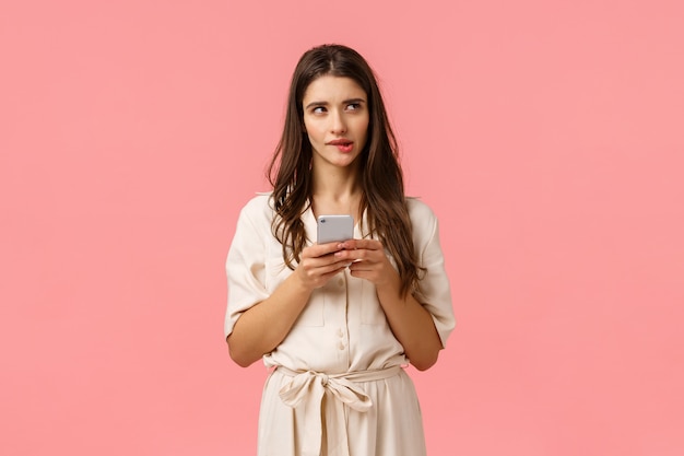 Photo curious and dreamy alluring woman shoppaholic thinking what order best, biting lip looking up imaging, holding phone, not sure sending risky message, standing pink wall thoughtful