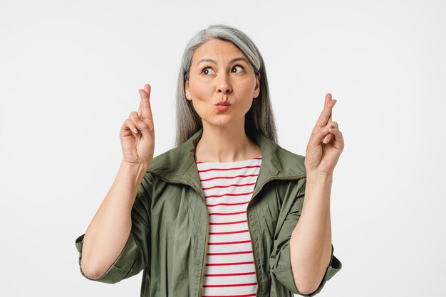 Curious dreamy mature middleaged caucasian woman with grey hair making a wish crossing fingers for good luck isolated in white background Dreams come true