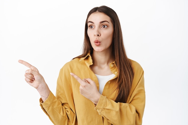 Curious female customer seeing cool new promo deal, pointing and looking aside at copy space left, showing banner logo, standing in casual clothes against white wall