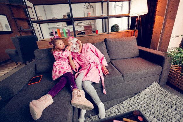 Curious girl. Curious preschool girl having much fun while putting sheet mask on granny sitting on sofa near her