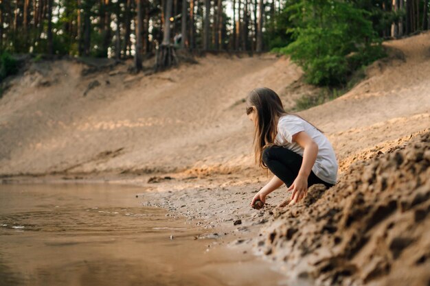 Фото Любопытная девушка с коричневыми волосами приседает на песке возле реки в лесу и собирается взять песчинку вода отражает ребенка
