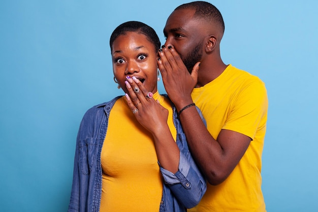 Curious girlfriend in disbelief listening to gossip told by boyfriend over blue background. Man wihispering secret and spreading rumour, keeping conversation confidential. Shocked people