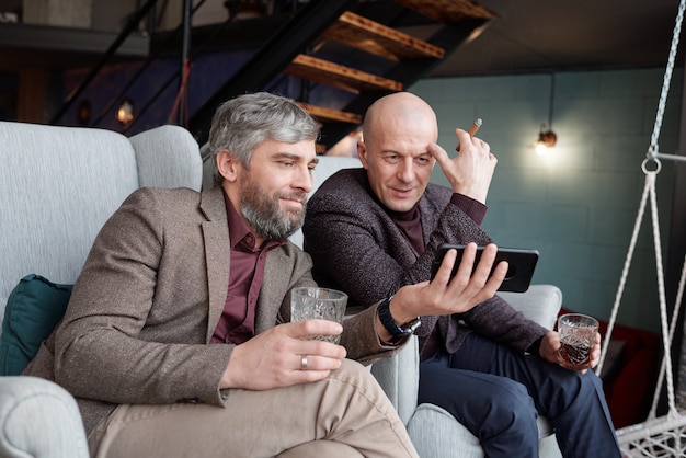 Curiosi bei uomini di mezza età che bevono whisky e guardano video online tramite smartphone