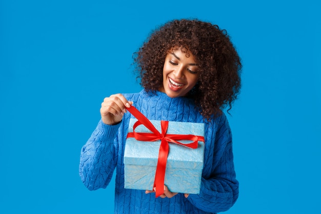 Curiosa e felice, sorridente donna afro-americana, ragazza di b-day in maglione invernale, tirando il nodo presente per scartare il regalo e vedere cosa dentro, festeggiare natale, vacanze di capodanno, sfondo blu