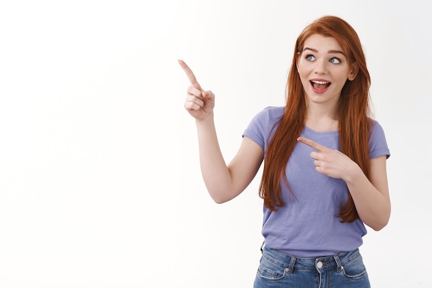 Curious joyful smiling redhead woman in purple t-shirt, standing intrigued or entertined, pointing looking upper left corner amused, see interesting advertisement, offer visit corporate site