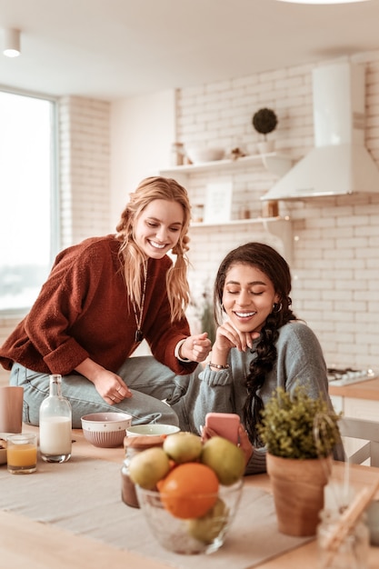 Curious light-haired girlfriend. Beaming attractive girls spending great time together while checking social media and eating breakfast