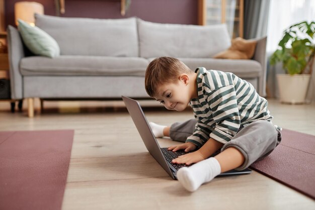집에서 바닥에 앉아 노트북을 사용하여 다운 증후군을 가진 호기심 어린 소년