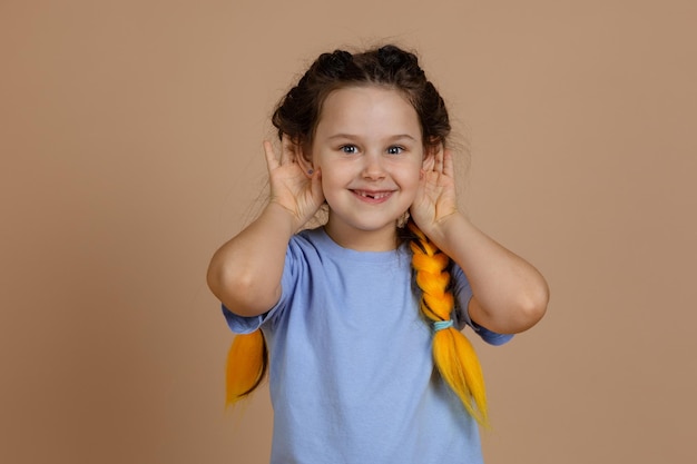 Photo curious small caucasian girl with kanekalon braids smiling with missing tooth holding ears to overhear something interesting looking at camera on beige background