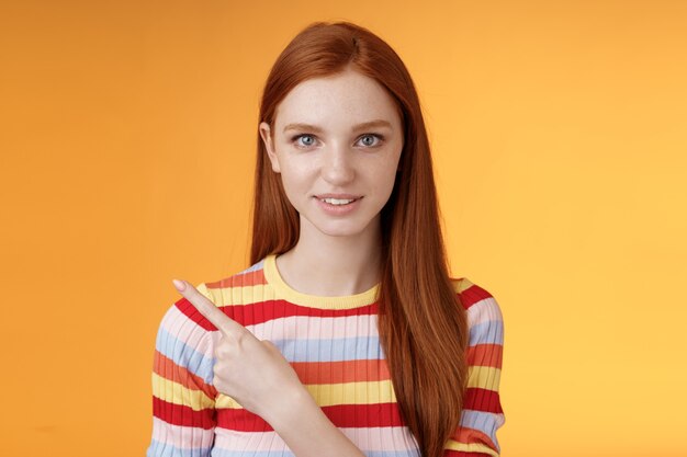 Curious smart female redhead coworker asking friend who was that pointing left index finger look camera thoughtful questioned wanna know answer smiling intrigued standing orange background