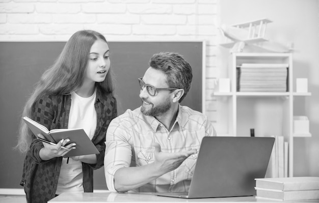 Curious teen girl and teacher man in high school with workbook\
and pc at blackboard parenthood